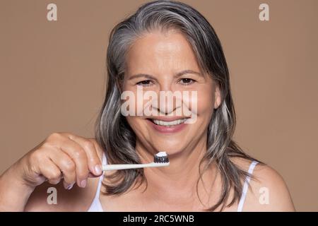 Joyeuse vieille dame caucasienne aux cheveux gris se brosse les dents, isolée sur fond brun, studio Banque D'Images