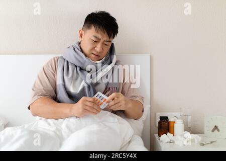 Homme chinois malsain assis dans son lit, prenant des pilules Banque D'Images