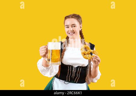 Jeune femme dans des vêtements traditionnels allemands avec de la bière et du bretzel sur fond jaune Banque D'Images