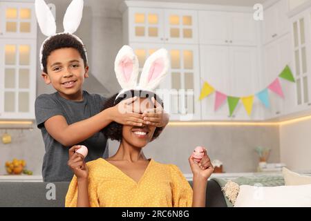 Mignon fils afro-américain couvrant les yeux de sa mère avec les mains alors qu'elle tient des oeufs de Pâques dans la cuisine Banque D'Images