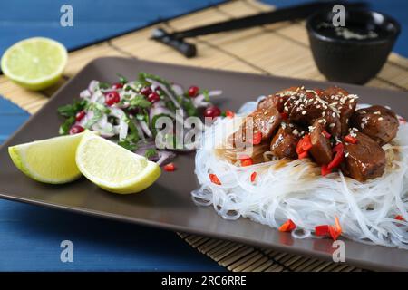 Morceaux de poulet sauce soja avec nouilles, salade et citron vert servis sur table bleue, gros plan Banque D'Images