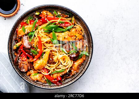 Faire sauter les nouilles avec les tranches de poulet, le paprika rouge, les champignons, la ciboulette, la sauce soja et les graines de sésame dans un bol en céramique. Plat de cuisine asiatique. Dos de table blanc Banque D'Images