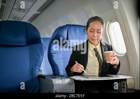 Une femme d'affaires asiatique âgée heureuse utilisant sa tablette numérique et sirotant un café pendant le vol pour son voyage d'affaires. Banque D'Images