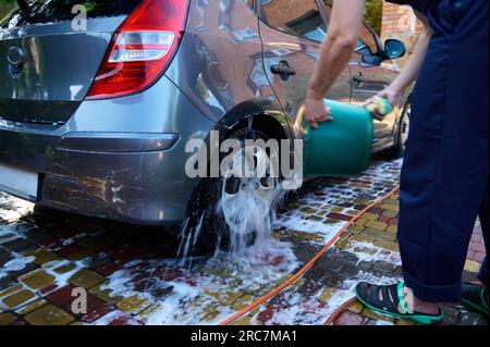 Mise au point sélective. Vue arrière d'un homme lavant manuellement sa voiture dans l'arrière-cour, versant de l'eau du seau sur la roue. Concept de lave-auto manuel. Peopl Banque D'Images