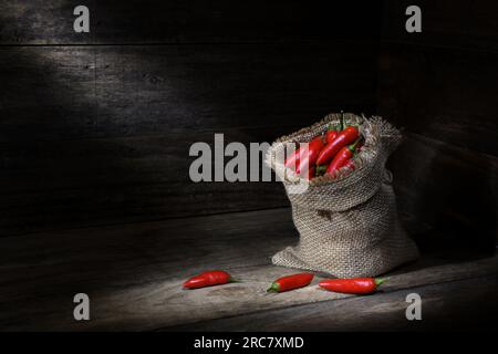 Une nature morte d'un sac hessien plein de piments rouges dans une boîte en bois rustique avec des piscines d'éclairage d'humeur sombre doux avec espace de copie Banque D'Images