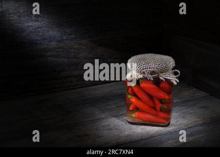 Une nature morte de piments rouges marinés faits maison dans un bocal en verre dans une boîte en bois rustique avec des piscines d'éclairage d'ambiance douce et sombre avec espace de copie Banque D'Images