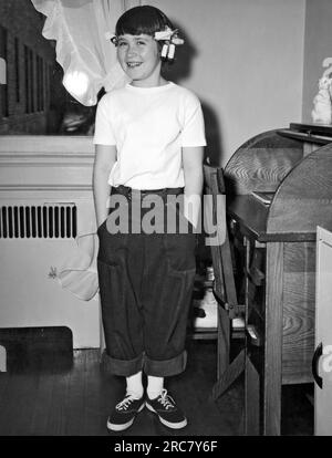 New York, New York : c. 1940 Une jeune fille souriante portant un Jean roulé, des chaussures de tennis et des bigoudis dans les cheveux se tient près de son bureau dans sa chambre. Banque D'Images