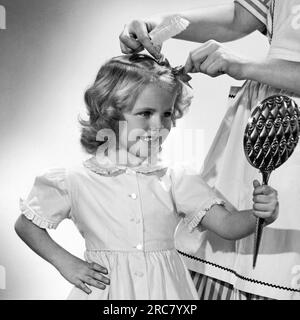 États utilisés : c. 1950. Une jeune fille regarde attentivement dans un miroir tandis que sa mère lui attache un arc dans les cheveux. Banque D'Images