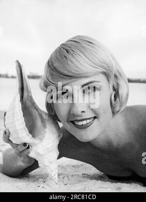 États-Unis : c. 1962. Une jolie femme blonde est allongée sur la plage avec une coquille de mer à l'oreille. Banque D'Images