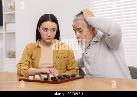 Jouer aux dames. Homme senior émotionnel apprenant jeune femme à table dans la salle Banque D'Images