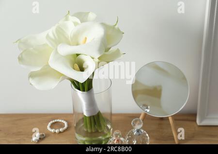 Belles fleurs de lys calla dans un vase en verre, bouteilles avec des parfums, des bijoux et un miroir sur une table en bois Banque D'Images