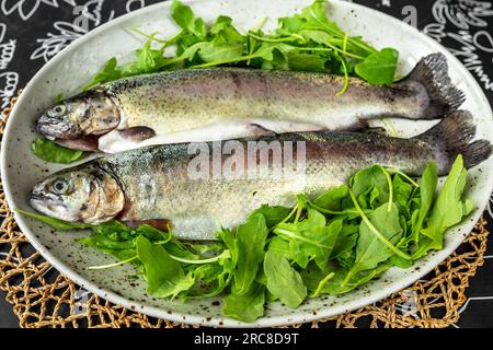 Deux truites arc-en-ciel crues sur assiette garnies de nombreuses feuilles vertes fraîches, closeup. Préparation de repas. Banque D'Images