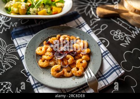 Crevettes grillées avec piment et oignon dans une assiette grise avec fourchette sur une serviette, pain et bol avec salade sur une table noire. Banque D'Images