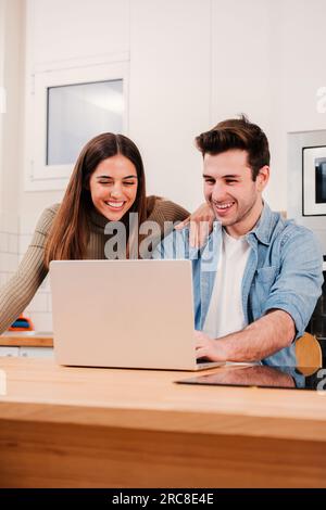 Portrait vertical de jeune couple naviguant sur Internet à la cuisine à domicile à l'aide d'un ordinateur portable. Heureux mari et femme planifiant les vacances avec un ordinateur parlant et souriant ensemble, regardant des vidéos drôles. Photo de haute qualité Banque D'Images