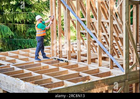 Menuisier construisant une maison à ossature en bois de deux étages près de la forêt. Homme barbu martelant des clous dans la structure tout en portant un casque de protection. Concept de construction écologique moderne. Banque D'Images