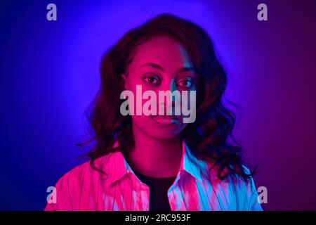 Portrait rapproché de belle, jeune, femme africaine regardant la caméra, posant sur fond bleu dégradé de studio violet dans la lumière néon Banque D'Images