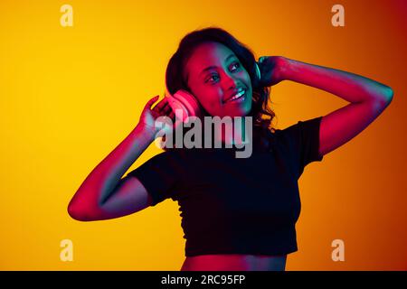 Portrait de jeune belle femme africaine écoutant de la musique dans des écouteurs sur fond de studio jaune dans la lumière néon. Plaisir Banque D'Images