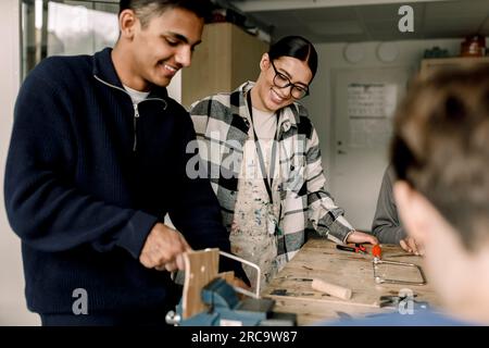 Heureuse enseignante féminine debout avec l'étudiant masculin coupant du bois pendant la classe de menuiserie Banque D'Images