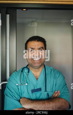 Portrait d'un médecin mûr joyeux debout avec les bras croisés devant la porte de l'hôpital Banque D'Images