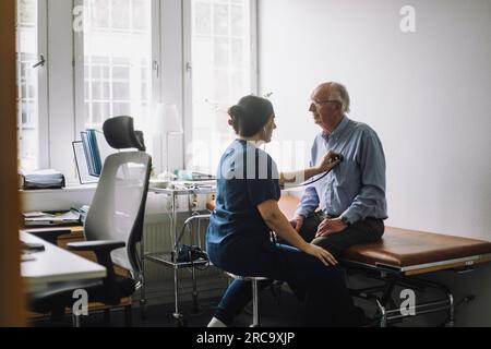 Travailleuse de santé examinant une patiente âgée assise sur son lit dans une clinique Banque D'Images