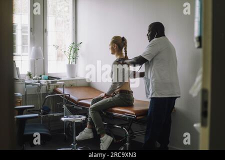 Vue latérale du médecin masculin mature examinant la patiente assise sur le lit en clinique Banque D'Images