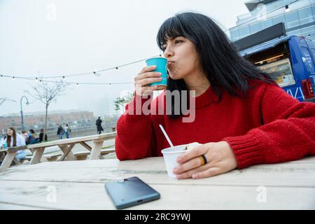jeune femme latine d'origine vénézuélienne, portant un chandail rouge, assise dehors pose le téléphone sur la table pour profiter d'une tasse de café avec un ic Banque D'Images