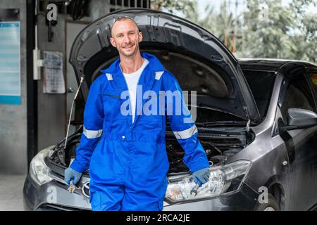 Heureux mécanicien de voiture souriant en combinaison bleue debout devant une voiture avec son capot s'ouvre Banque D'Images