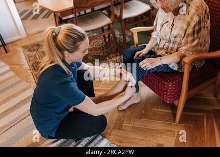 Vue à grand angle d'une travailleuse de santé aidant une femme âgée portant des chaussettes à la maison Banque D'Images