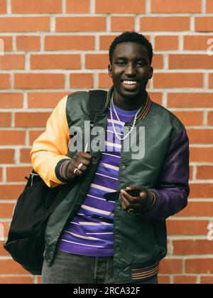 Portrait de jeune homme heureux tenant des lunettes de soleil tout en se tenant debout contre le mur de briques Banque D'Images