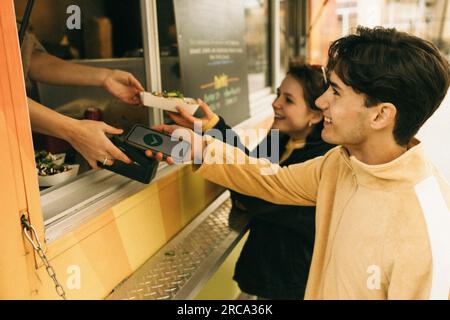 Jeune homme souriant faisant le paiement en ligne tout en achetant de la nourriture du stand de concession Banque D'Images