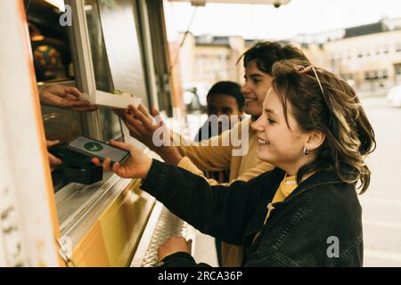 Jeune femme souriante faisant le paiement en ligne par téléphone intelligent tout en achetant de la nourriture du stand de concession Banque D'Images