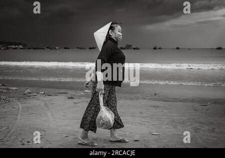 Une femme vietnamienne d'âge moyen portant un chapeau conique traditionnel porte un sac de fruits de mer sur la plage My Khe, Danang, Vietnam. Banque D'Images