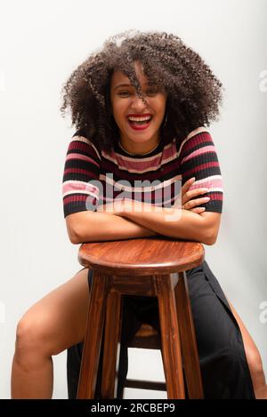 Portrait d'une belle jeune femme souriante et gaie avec des cheveux bouclés assis sur un tabouret en bois. Isolé sur fond gris clair. Banque D'Images