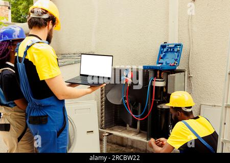 Électricien qualifié nettoyant et lubrifiant le compresseur interne du climatiseur extérieur, vérifiant le niveau de fréon tandis que le collègue afro-américain vérifie le calendrier de maintenance sur l'ordinateur portable Banque D'Images