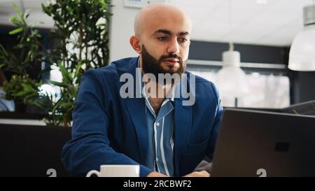 Homme d'affaires résolvant des problèmes de démarrage avec ordinateur portable et documents archivés, regardant le rapport de statistiques annuel pour trouver le problème. Jeune analyste examinant des articles, plan stratégique exécutif. Banque D'Images