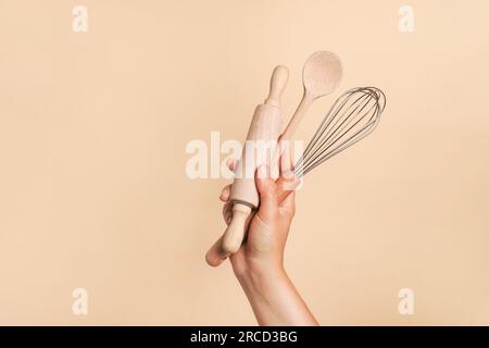 Main féminine tenant des ustensiles de cuisine pour la nourriture et la boulangerie sur fond beige. fouet, rouleau à pâtisserie et cuillère en bois Banque D'Images