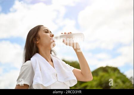 Une jeune femme asiatique en bonne santé et belle en vêtements de sport boit de l'eau d'une bouteille, a soif, se repose après une longue course sur une journée ensoleillée à un Banque D'Images