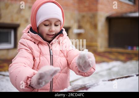 Portrait authentique d'une adorable petite fille en veste rose chaude en duvet, ramasser la neige d'une surface couverte de neige, faire des boules de neige, jouer o Banque D'Images