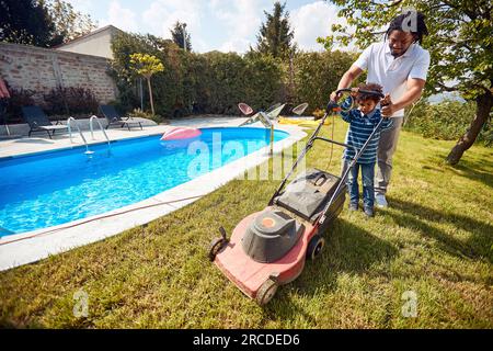 Moment tendre comme un père guide son jeune fils dans l'art de tondre la pelouse, juste à côté de l'oasis tranquille de la piscine. Banque D'Images