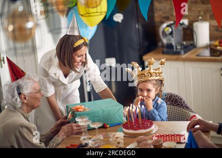 Heureuse petite fille faisant un vœu avant de souffler des bougies sur le gâteau, assis à la table de la cuisine avec sa famille autour, mère et grands-parents, l'acclamant Banque D'Images