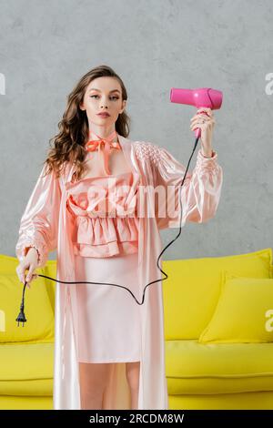 photographie de concept, femme agissant comme une poupée, belle femme au foyer en robe de soie rose tenant sèche-cheveux et prise, debout près de l'entraîneur jaune dans moderne Banque D'Images