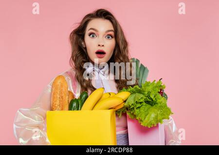 concept de femme au foyer, jeune femme choquée portant des sacs d'épicerie avec des légumes et des bananes, modèle avec des cheveux ondulés sur fond rose, photogra conceptuel Banque D'Images