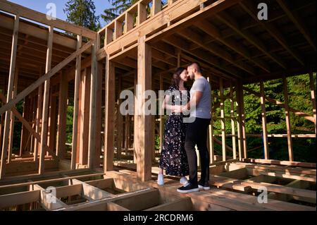 Homme et femme inspectant leur future maison de cadre en bois nichée près de la forêt. Jeune couple sur le chantier en début de matinée. Concept de construction écologique contemporaine. Banque D'Images