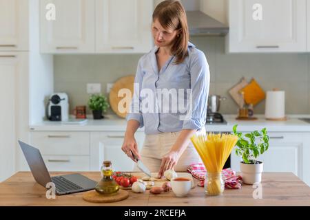 Une jeune femme cuisine à la maison dans la cuisine, elle regarde une recette vidéo sur un ordinateur portable et travaille avec la pâte sur la table, elle coupe l'ail. Cui italien Banque D'Images