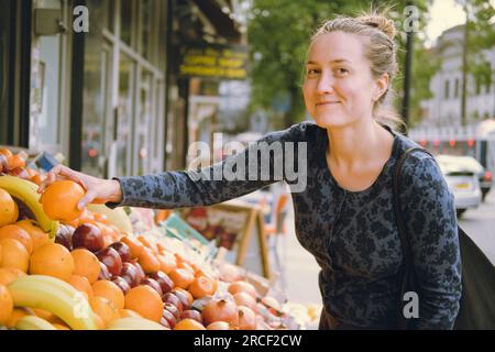 Femme jeune-adulte souriante cueillant une orange avec sa main droite magasinant dans un magasin d'alimentation, choisissant ce qu'il faut acheter au stand de fruits sur un marché de rue, l Banque D'Images