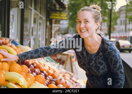 Jeune adulte femelle riant et touchant des oranges avec sa main droite lors de ses achats dans un magasin d'alimentation, choisissant ce qu'il faut acheter au stand de fruits sur une stree Banque D'Images