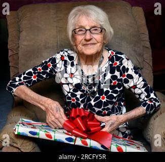 Une grand-mère de 100 ans s'assoit dans sa chaise préférée et ouvre un paquet coloré le matin de Noël.Elle regarde avec un sourire et scintillent en elle Banque D'Images