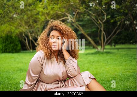 Gros plan portrait de Jeune belle femme afro-américaine profite d'une promenade dans le parc vert public d'été. Une femme heureuse plus taille portant une robe rose soie assise sur l'herbe Banque D'Images