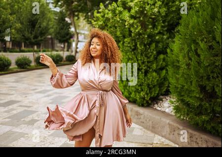 Jeune belle femme afro-américaine profite d'une promenade dans le parc vert public d'été. Une femme heureuse plus taille portant la robe rose soie sourit se promène dans les rues de la ville Banque D'Images