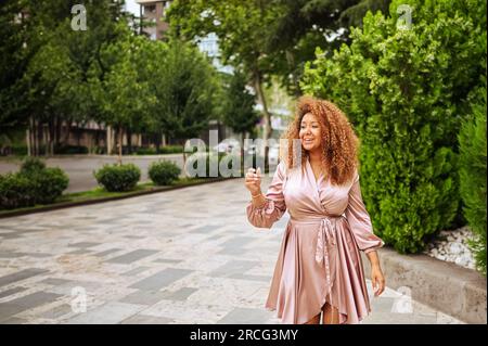 Jeune belle femme afro-américaine profite d'une promenade dans le parc vert public d'été. Une femme heureuse plus taille portant la robe rose soie sourit se promène dans les rues de la ville Banque D'Images